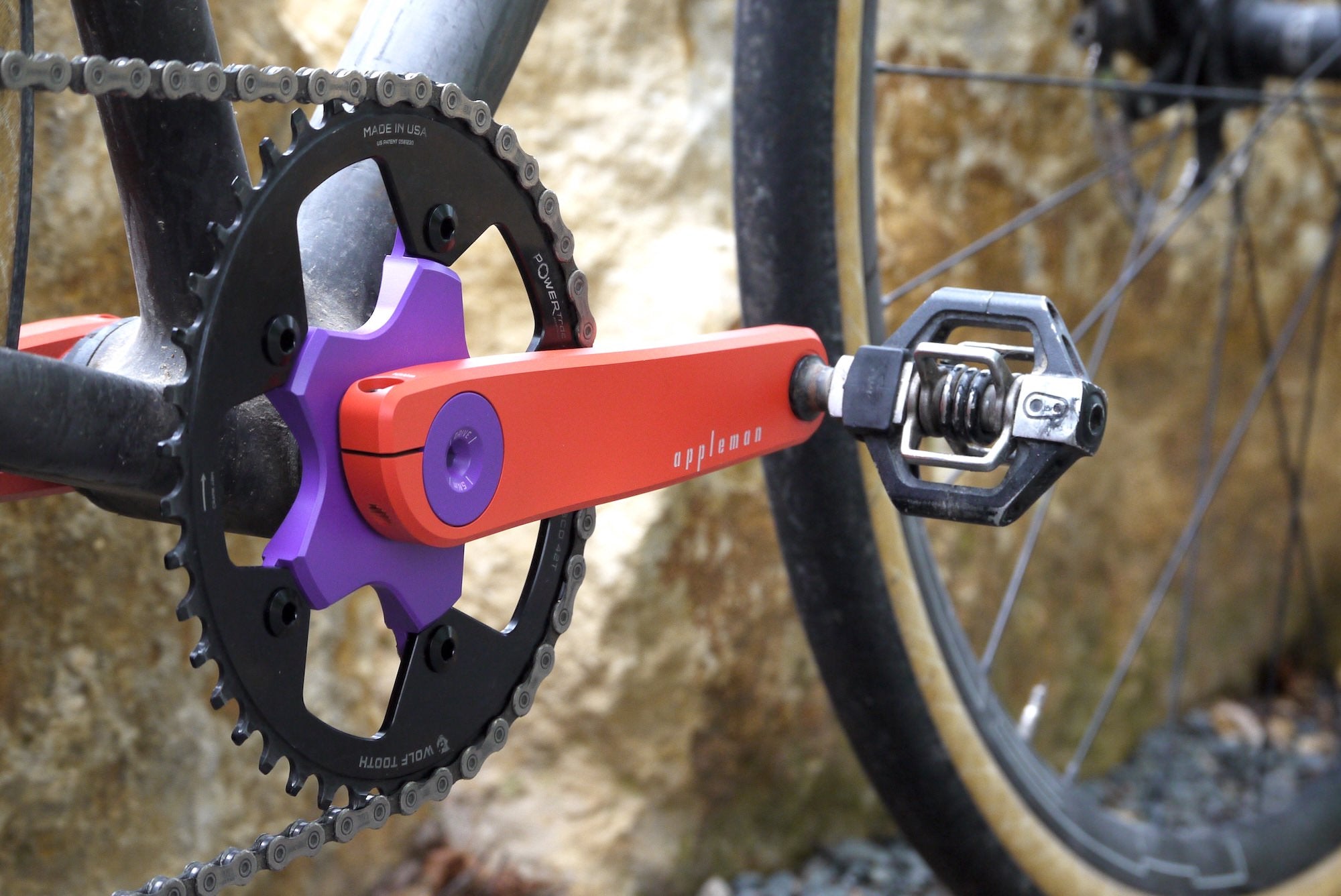 Close-up of a bicycle crankset with a bright orange crank arm, purple spindle bolt, and a black chainring, attached to a carbon fiber bicycle